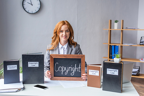 Smiling lawyer showing chalkboard with copyright lettering near books, while sitting at workplace with documents
