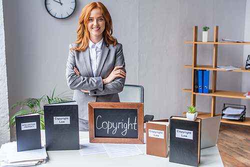 Smiling lawyer with crossed arms standing near chalkboard with copyright lettering near books on table with pile of documents