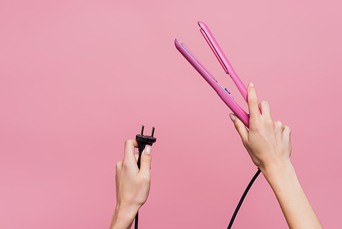 cropped view of woman holding hair straightener in hands isolated on pink