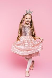 full length of cheerful little girl in dress and crown posing on pink