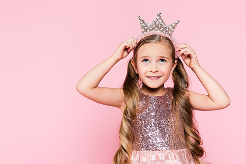 cheerful little girl in dress adjusting crown isolated on pink