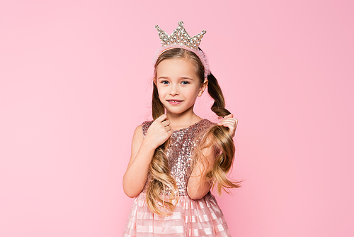 cheerful little girl in dress and crown adjusting hair isolated on pink