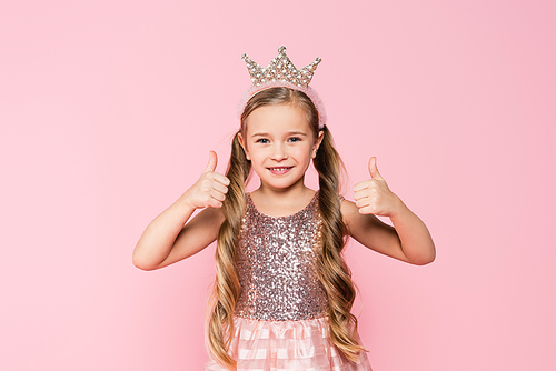 cheerful little girl in dress and crown showing thumbs up isolated on pink