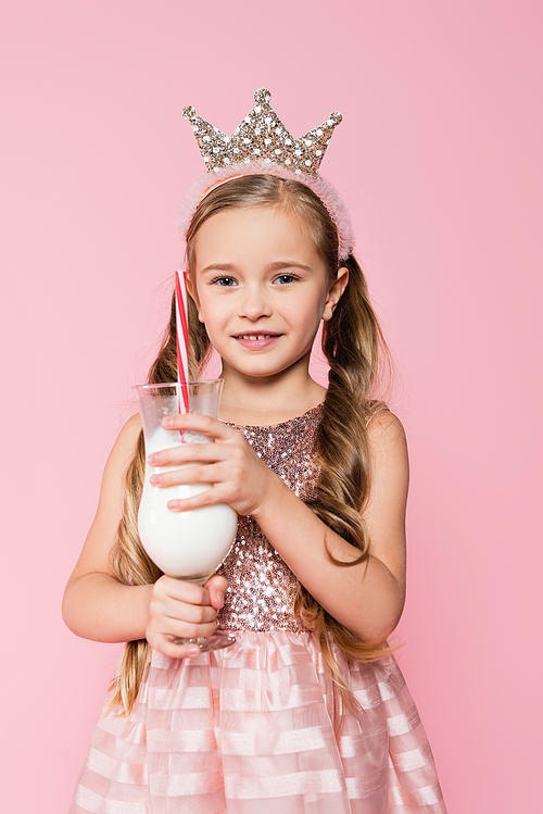 cheerful little girl in dress and crown holding glass with milkshake isolated on pink