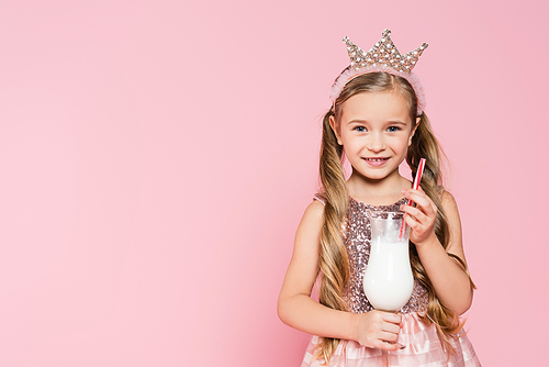 happy little girl in dress and crown holding glass with milkshake isolated on pink