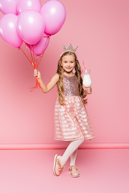 full length of cheerful little girl in dress and crown holding glass with milkshake and balloons on pink