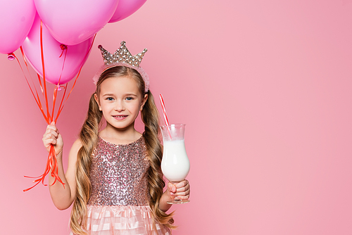 cheerful little girl in dress and crown holding glass with milkshake and balloons isolated on pink