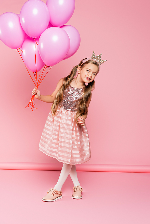 full length of cheerful little girl in dress and crown holding balloons on pink