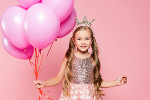 cheerful little girl in dress and crown holding balloons isolated on pink