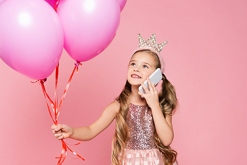 cheerful little girl in dress and crown holding balloons and talking on smartphone isolated on pink