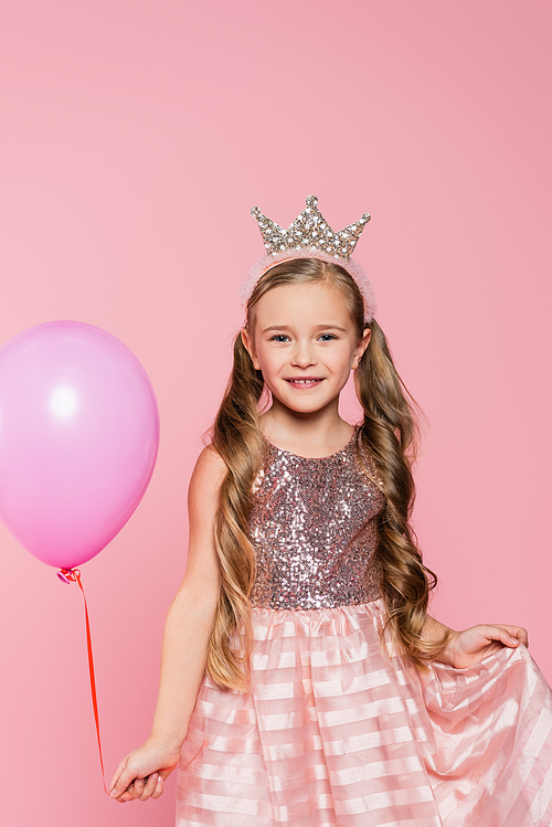joyful little girl in dress and crown holding balloon isolated on pink