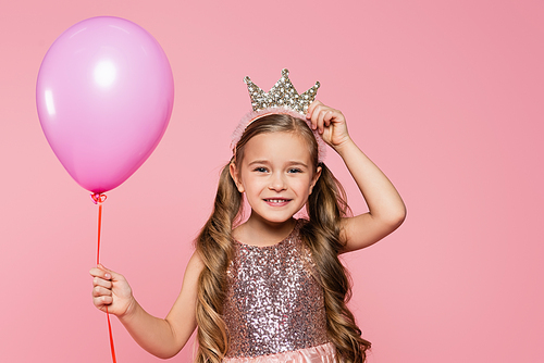 joyful little girl in dress adjusting crown and holding balloon isolated on pink