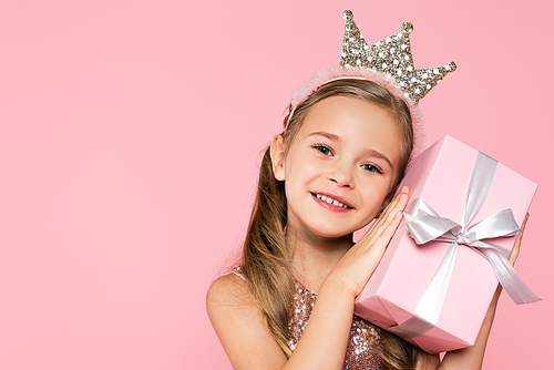 cheerful little girl in crown holding wrapped present isolated on pink