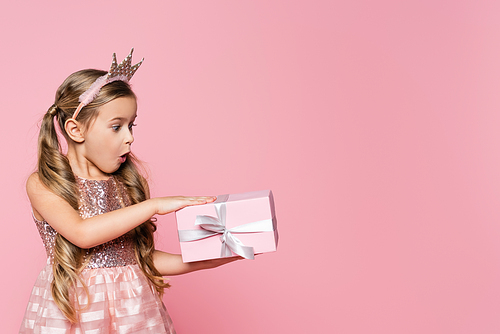 surprised little girl in crown looking at wrapped present isolated on pink