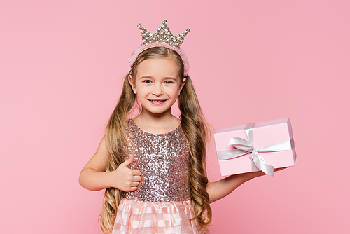 joyful little girl in crown holding wrapped present showing thumb up isolated on pink