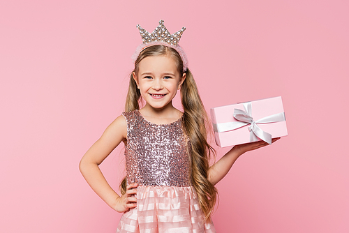 joyful little girl in crown holding wrapped present and standing with hand on hip isolated on pink