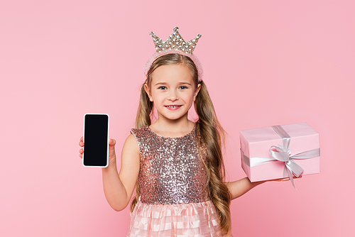 joyful little girl in crown holding wrapped present and smartphone with blank screen  isolated on pink