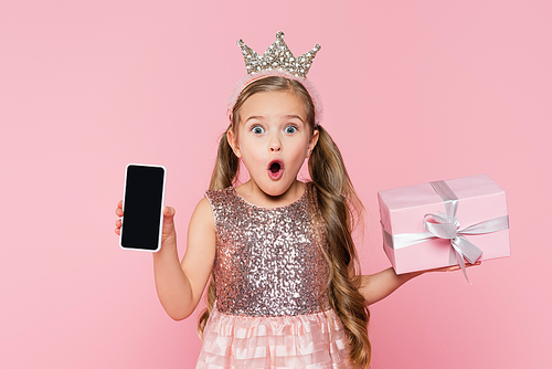 shocked little girl in crown holding wrapped present and smartphone with blank screen  isolated on pink