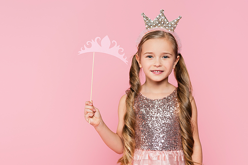 cheerful little girl in dress holding carton crown on stick isolated on pink