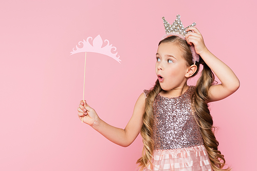 shocked little girl in dress holding paper crown on stick isolated on pink