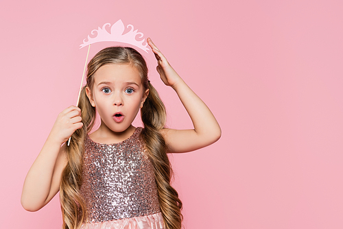 surprised little girl in dress holding paper crown on stick above head isolated on pink