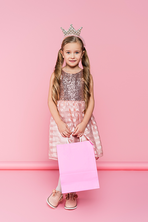 full length of cheerful little girl in dress and crown holding shopping bag on pink