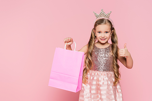 cheerful little girl in dress and crown holding shopping bag and showing thumb up isolated on pink