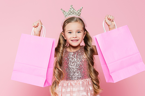 cheerful little girl in dress and crown holding shopping bags isolated on pink