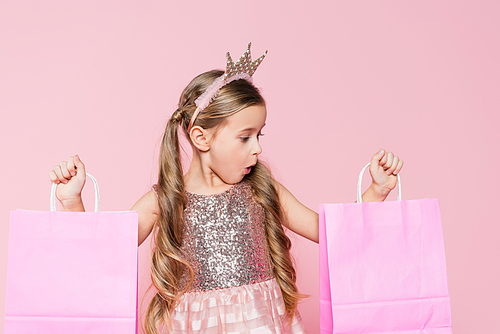 shocked little girl in crown holding shopping bags isolated on pink