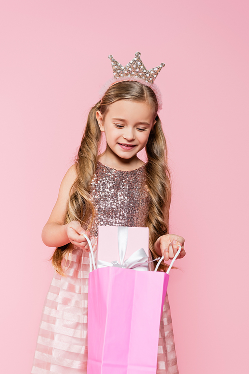 little girl in dress and crown holding shopping bag with present isolated on pink