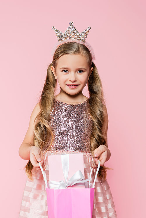 happy little girl in dress and crown holding shopping bag with present isolated on pink