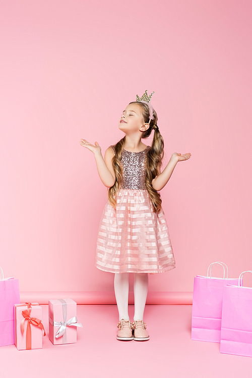 full length of pleased little girl in crown with closed eyes standing near presents and shopping bags on pink