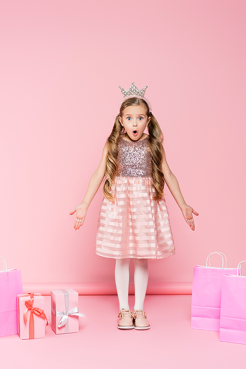 full length of shocked little girl in crown standing near presents and shopping bags on pink