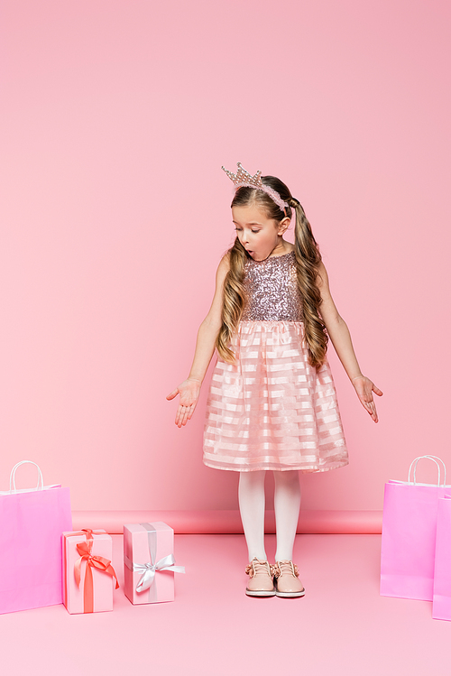 full length of surprised little girl in crown standing near presents and shopping bags on pink