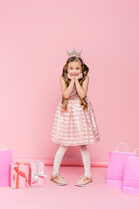 full length of excited little girl in crown standing near presents and shopping bags on pink