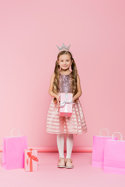 full length of smiling little girl in crown holding present near shopping bags on pink