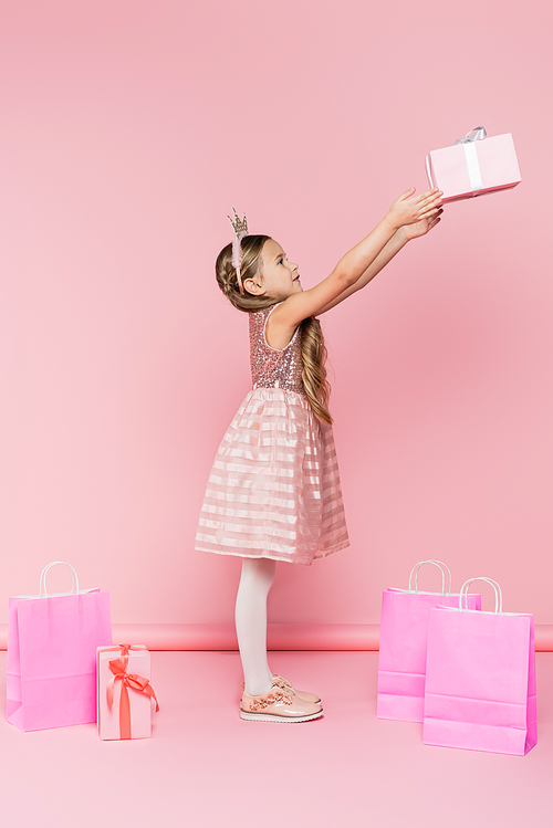 full length of happy little girl in crown holding present near shopping bags on pink