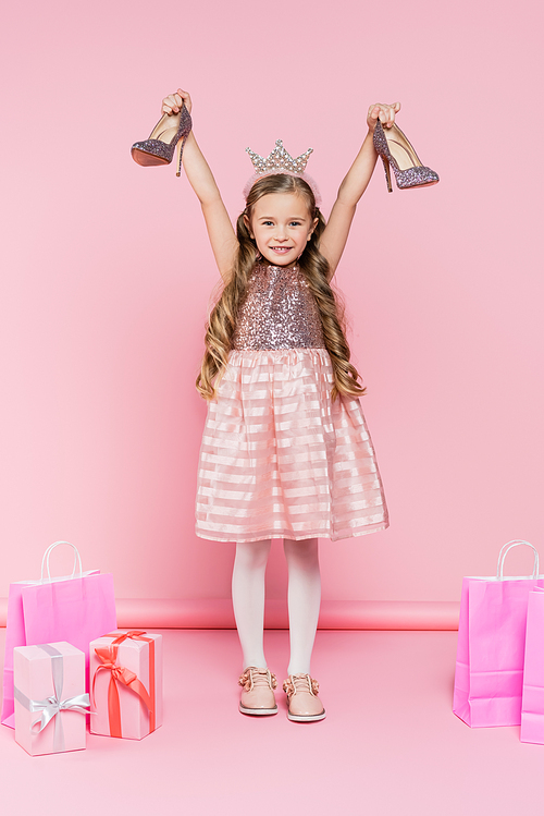 full length of cheerful little girl in crown holding heels near presents and shopping bags on pink