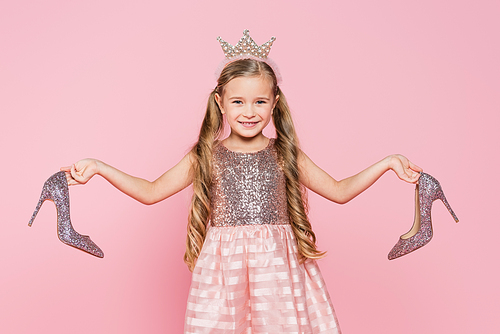 cheerful little girl in crown and dress holding heels isolated on pink