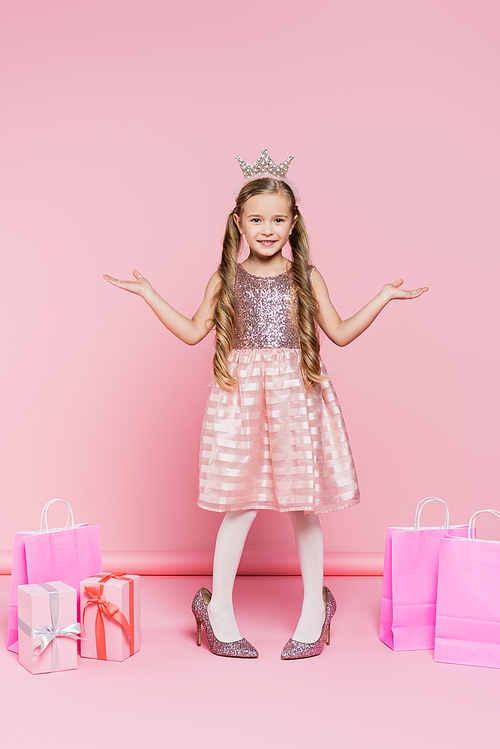 full length of happy little girl in crown standing on heels near presents and shopping bags on pink