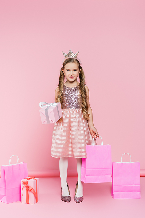 full length of happy little girl in crown standing on heels and holding present and shopping bag on pink