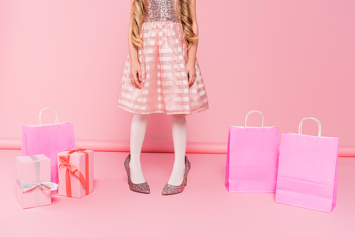 cropped view of little girl in dress standing on heels near presents and shopping bags on pink