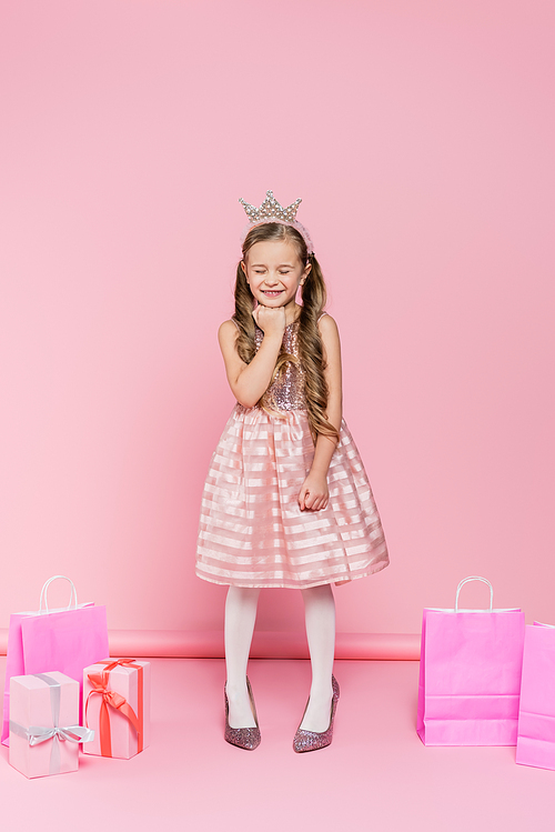 full length of happy little girl in crown standing on heels near presents and shopping bags on pink