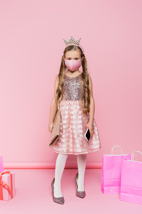 full length of little girl in crown and medical mask standing on heels, holding smartphone and bag near present and shopping bags on pink