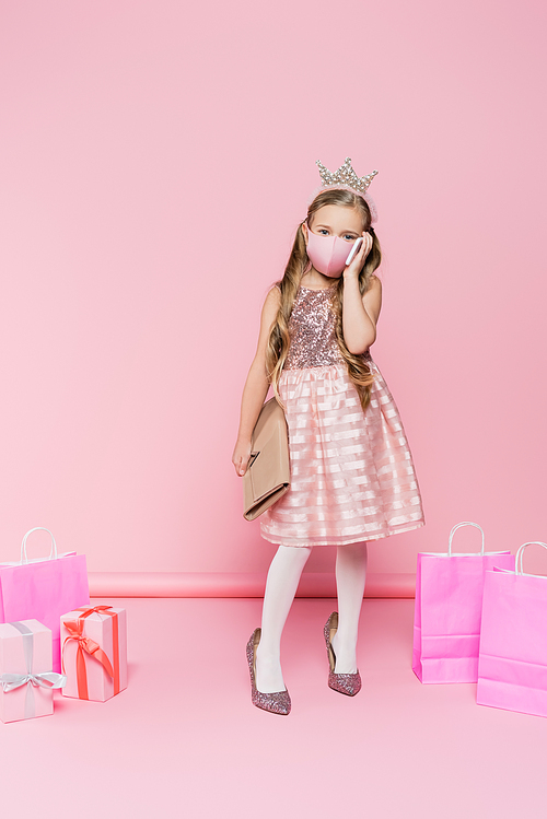 full length of girl in crown and medical mask standing on heels, talking on smartphone while holding bag near presents and shopping bags on pink