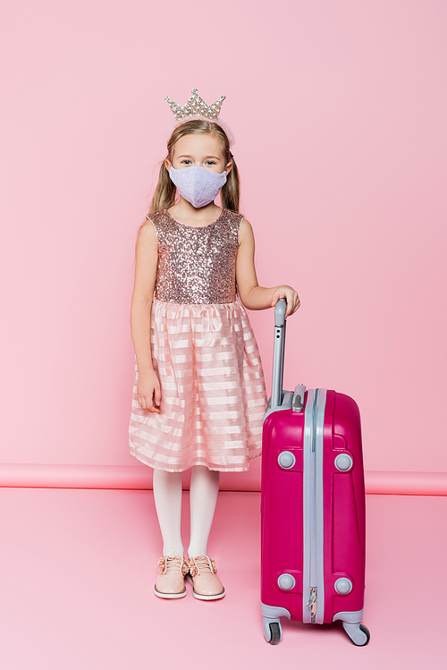 full length of little girl in crown and medical mask standing near baggage on pink