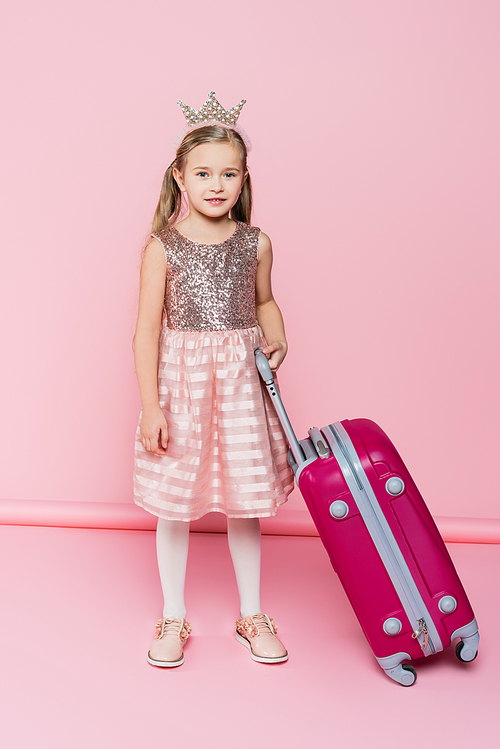 full length of happy little girl in crown and dress standing near baggage on pink