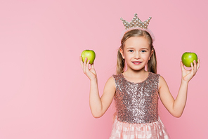 happy little girl in crown holding green apples isolated on pink