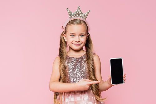 happy little girl in crown pointing with finger at smartphone with blank screen isolated on pink
