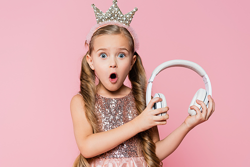 shocked little girl in crown holding wireless headphones isolated on pink